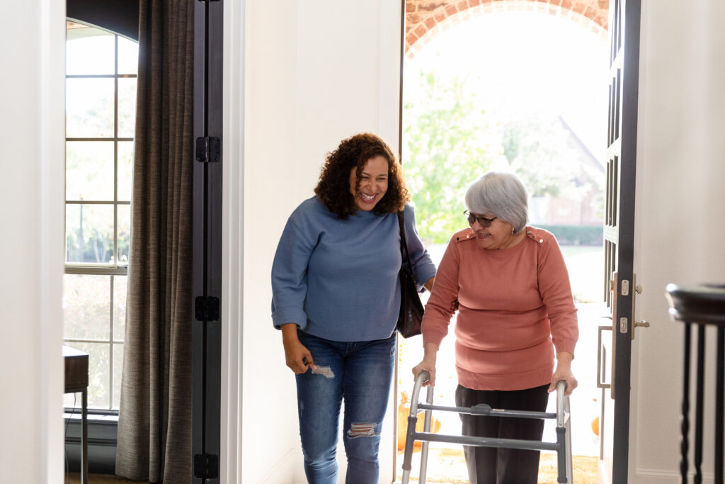 Two women enter the front door of a home. One is elderly and uses a walking frame. The other is middle-aged and is assisting the older woman.